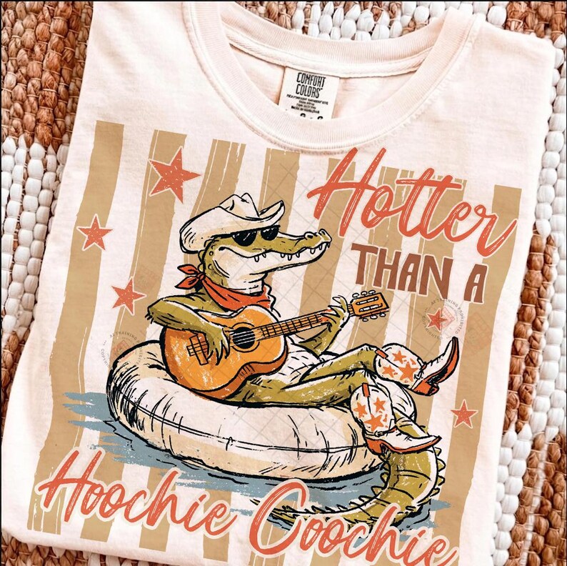 May include: Cream-colored t-shirt with a graphic of an alligator wearing a cowboy hat, sunglasses, and boots, playing a guitar while floating on an inner tube. The text reads "Hotter Than A Hoochie Coochie."
