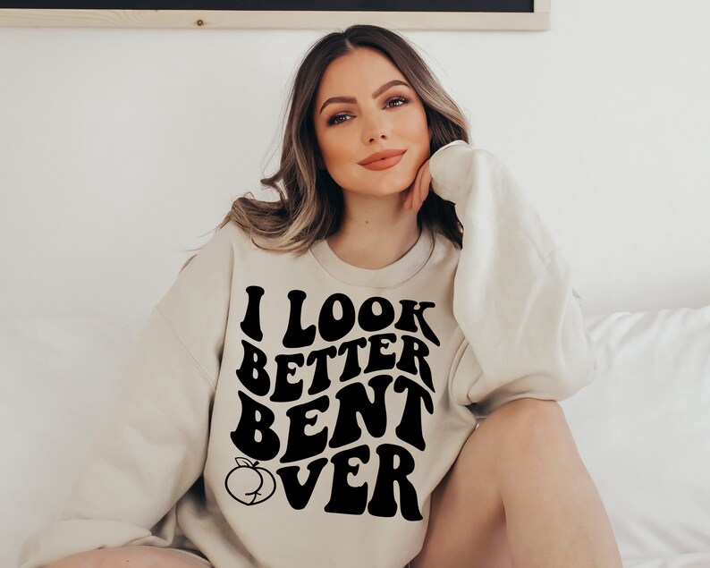 May include: A cream-colored sweatshirt with black text that reads "I look better bent over" in a retro font. A small peach graphic is next to the text.