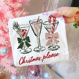 May include: White t-shirt featuring three martini glasses with festive holiday decorations. The glasses are adorned with bows and garnishes, including cherries and a candy cane. The words "Christmas please" are written in red script.