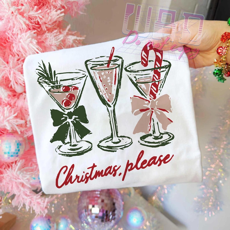 May include: White t-shirt featuring three martini glasses with festive holiday decorations. The glasses are adorned with bows and garnishes, including cherries and a candy cane. The words "Christmas please" are written in red script.