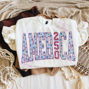 May include: White crewneck sweatshirt with the word "AMERICA 250" in blue and red, with a star pattern inside the letters. The sweatshirt is folded on top of other clothing items.
