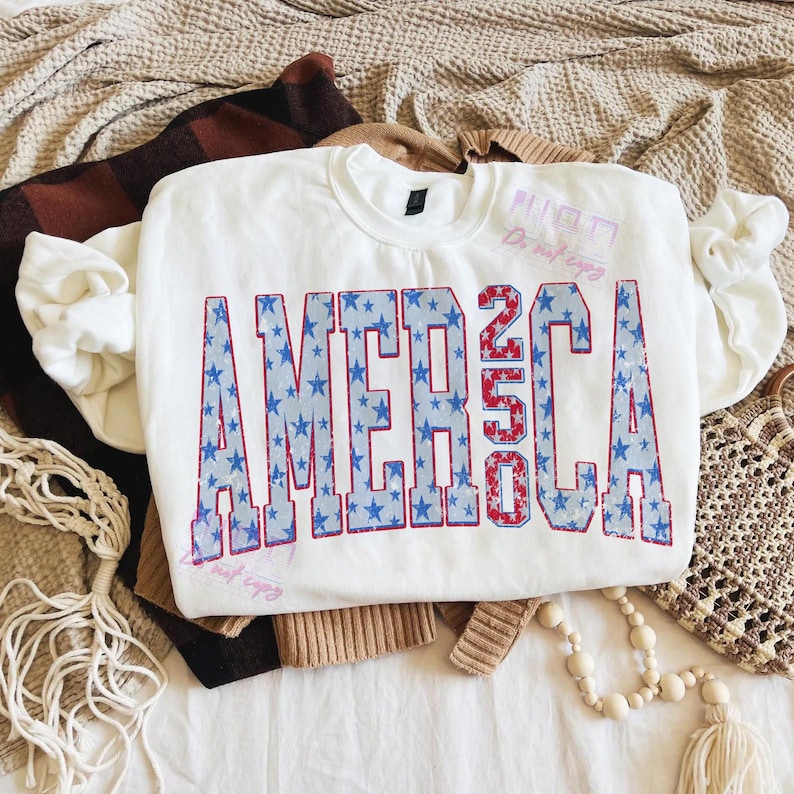 May include: White crewneck sweatshirt with the word "AMERICA 250" in blue and red, with a star pattern inside the letters. The sweatshirt is folded on top of other clothing items.
