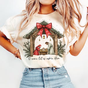 May include: Cream-colored t-shirt featuring a Nativity scene with a red bow, snow, and the text "O come let us adore Him." The graphic depicts Mary, Joseph, and baby Jesus in a manger, under a wooden structure.