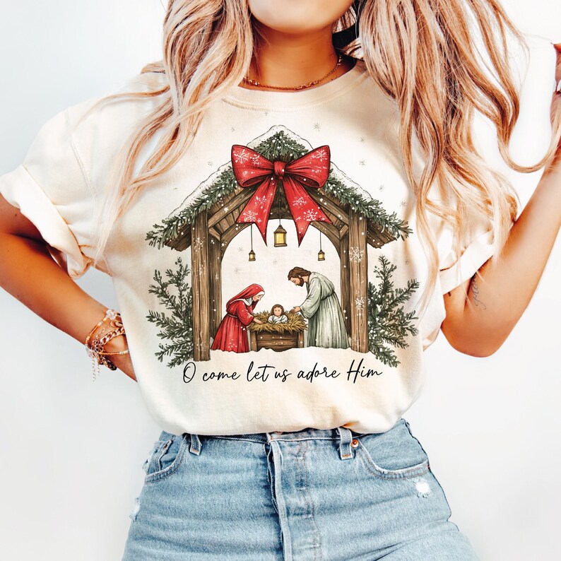 May include: Cream-colored t-shirt featuring a Nativity scene with a red bow, snow, and the text "O come let us adore Him." The graphic depicts Mary, Joseph, and baby Jesus in a manger, under a wooden structure.