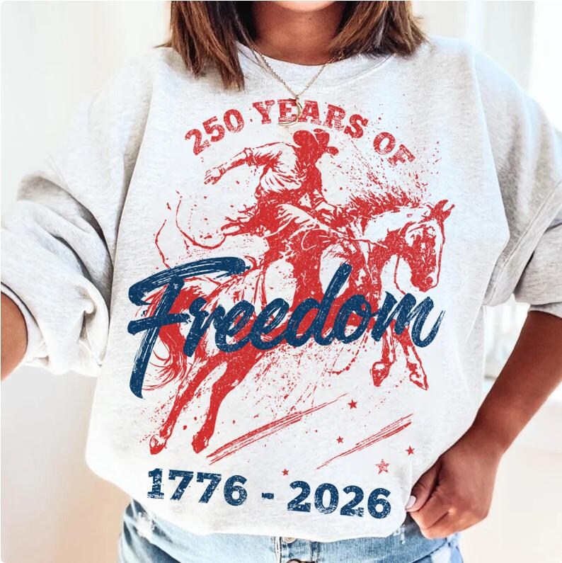 May include: Light gray sweatshirt with a red and blue graphic of a cowboy riding a horse. The text reads "250 Years of Freedom" above the image and "1776-2026" below. The design has a vintage, distressed look.
