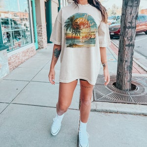 May include: A beige oversized t-shirt with a graphic print of a sunset scene with palm trees and the text "salt water sun shine".