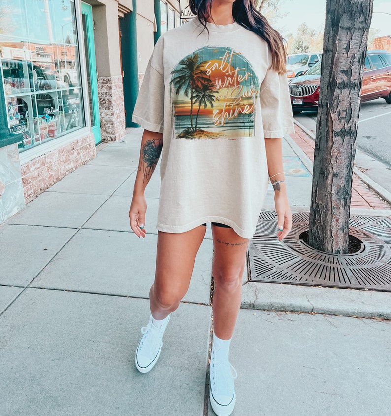 May include: A beige oversized t-shirt with a graphic print of a sunset scene with palm trees and the text "salt water sun shine".
