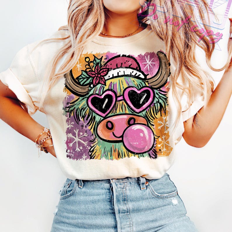 May include: Cream-colored t-shirt featuring a colorful cartoon Highland cow with heart-shaped sunglasses and a bubblegum bubble. The cow wears a Santa hat and has snowflakes in the background. The design is printed on a soft, casual tee.