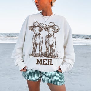 May include: White sweatshirt with a graphic of two goats wearing cowboy hats. The text "MEH." is printed below the goats.