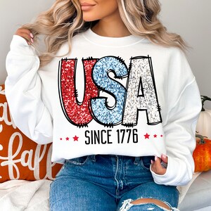 May include: White sweatshirt with a red, white, and blue glitter graphic of the word "USA" and the text "Since 1776".
