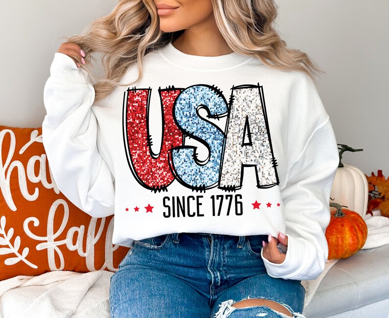 May include: White sweatshirt with a red, white, and blue glitter graphic of the word "USA" and the text "Since 1776".