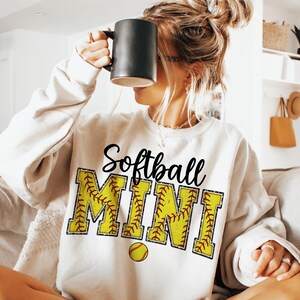 May include: White sweatshirt with the text "Softball Mini" in black lettering. The letters are outlined in a yellow and red softball pattern. A small yellow softball is below the text.