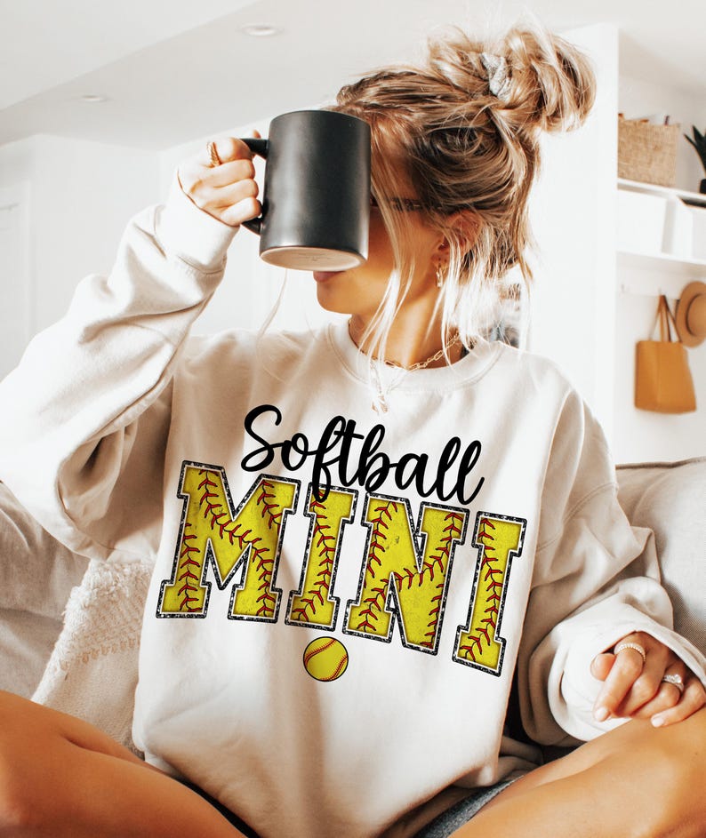 May include: White sweatshirt with the text "Softball Mini" in black lettering. The letters are outlined in a yellow and red softball pattern. A small yellow softball is below the text.