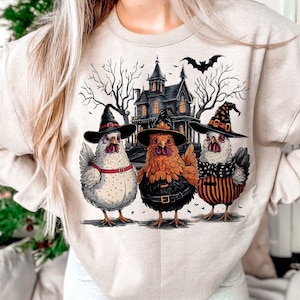 K&ouml;nnte beinhalten: Ein beiger Pullover mit einem Halloween-Motiv, das drei H&uuml;hner zeigt, die Hexenh&uuml;te tragen, und ein Spukhaus im Hintergrund.