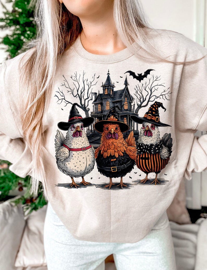 K&ouml;nnte beinhalten: Ein beiger Pullover mit einem Halloween-Motiv, das drei H&uuml;hner zeigt, die Hexenh&uuml;te tragen, und ein Spukhaus im Hintergrund.