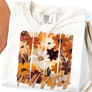 May include: Cream-colored t-shirt featuring a floral design in shades of orange, brown, and white, with the words "Fall Vibes" in an orange script. The shirt has a crew neck and short sleeves. A gold necklace is draped across the shirt.