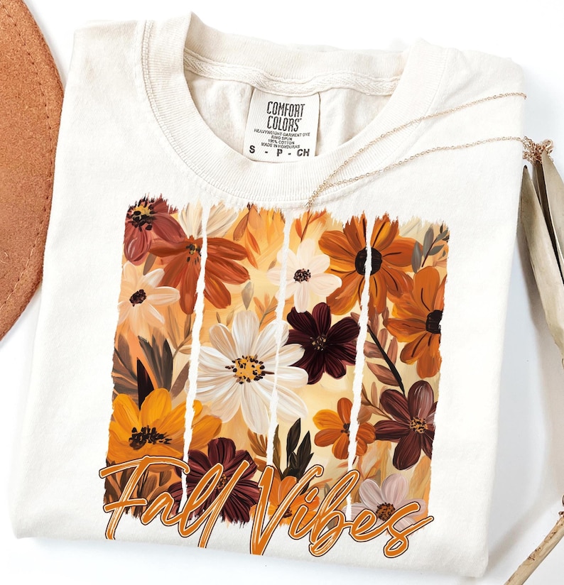 May include: Cream-colored t-shirt featuring a floral design in shades of orange, brown, and white, with the words "Fall Vibes" in an orange script. The shirt has a crew neck and short sleeves. A gold necklace is draped across the shirt.