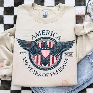 May include: A cream-colored sweatshirt featuring a patriotic design. The graphic includes a bald eagle with outstretched wings, the word "AMERICA," and the text "250 YEARS OF FREEDOM" with the years 1776 and 2026. The design is in red, white, and blue.