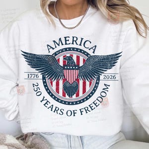 May include: White sweatshirt featuring a patriotic design with the word "AMERICA" above a circular emblem. The emblem includes a bald eagle with outstretched wings, a shield, and the text "250 YEARS OF FREEDOM" with the years 1776 and 2026.