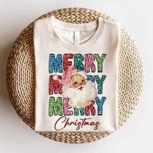 Op de afbeelding: Een wit T-shirt met een vintage kerstgrafiek. De grafiek toont de woorden "Merry Christmas" in kleurrijke, glinsterende letters en een cartoon kerstman met een rode muts en een witte baard.