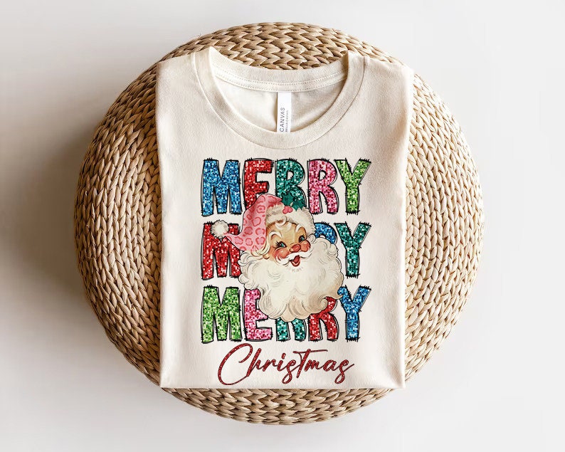 Op de afbeelding: Een wit T-shirt met een vintage kerstgrafiek. De grafiek toont de woorden "Merry Christmas" in kleurrijke, glinsterende letters en een cartoon kerstman met een rode muts en een witte baard.