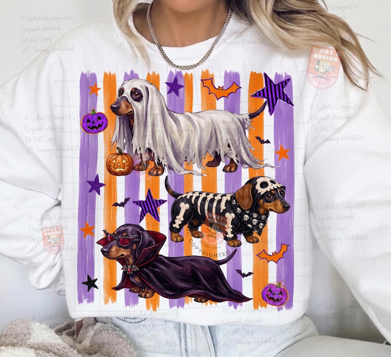 May include: A white crewneck sweatshirt featuring a graphic of three dachshunds in Halloween costumes. One dog wears a ghost sheet, one a skeleton suit, and one a vampire cape with sunglasses. The background has purple and orange vertical stripes.