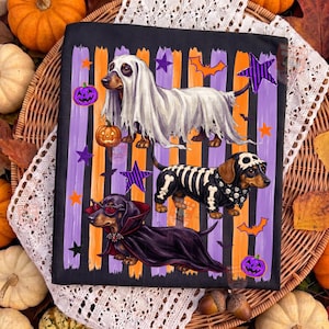 May include: A black t-shirt featuring three dachshunds in Halloween costumes: one as a ghost, one as a skeleton, and one as a vampire. The background has vertical purple and orange stripes with orange pumpkins, purple stars, and orange bats.