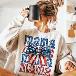 May include: Cream-colored sweatshirt with the word "Mama" in a patriotic design. The design features a large bow with a blue star pattern and red and white stripes. The sweatshirt is a casual style.