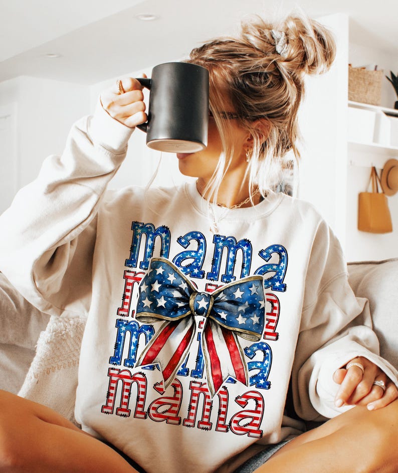 May include: Cream-colored sweatshirt with the word "Mama" in a patriotic design. The design features a large bow with a blue star pattern and red and white stripes. The sweatshirt is a casual style.