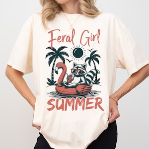 May include: A cream colored t-shirt with a graphic of a raccoon wearing sunglasses and a drink while floating on a flamingo pool float. The text "Feral Girl Summer" is printed on the shirt.