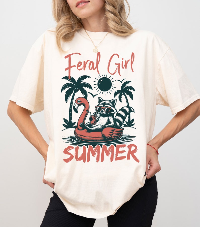 May include: A cream colored t-shirt with a graphic of a raccoon wearing sunglasses and a drink while floating on a flamingo pool float. The text "Feral Girl Summer" is printed on the shirt.
