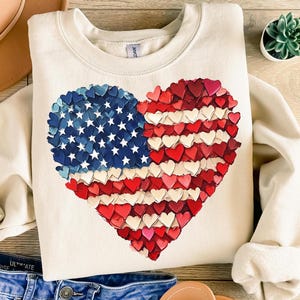 May include: A cream-colored sweatshirt with a large heart-shaped American flag design. The flag is made up of small red, white, and blue hearts.