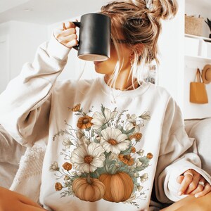 Pu&ograve; includere: Felpa color crema con un design floreale e zucca. La grafica include fiori bianchi, dettagli arancioni e due zucche. La felpa &egrave; a maniche lunghe e girocollo.