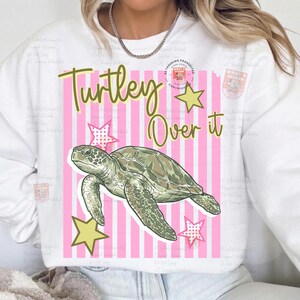May include: White sweatshirt with a graphic design featuring a green sea turtle, yellow stars, and the text "Turley Over it" on a pink striped background. The design is centered on the front of the sweatshirt.