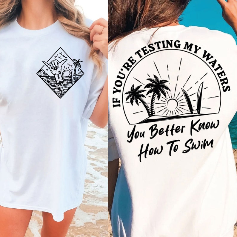May include: White t-shirt with a black graphic on the front of a hand giving a shaka sign with palm trees and waves. The back of the shirt has a black circle graphic with palm trees, a sun, and surfboards with the text "If you're testing my waters you better know how to swim".