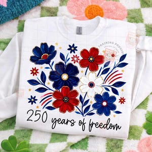May include: White sweatshirt featuring a floral design in red, white, and blue, with gold accents. The design includes the text "250 years of freedom" in a script font. The sweatshirt is a crew neck style.