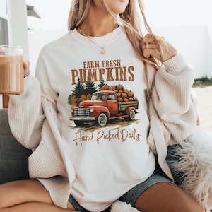 Op de afbeelding: Wit sweatshirt met een afbeelding van een rode pick-up truck vol met pompoenen. De tekst "Farm Fresh Pumpkins" en "Hand Picked Daily" staat op het shirt gedrukt.