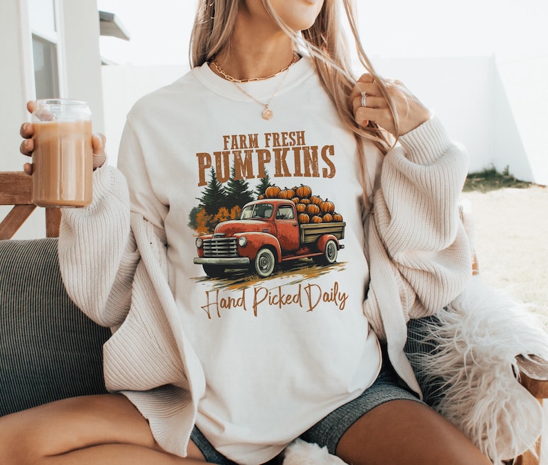 Op de afbeelding: Wit sweatshirt met een afbeelding van een rode pick-up truck vol met pompoenen. De tekst "Farm Fresh Pumpkins" en "Hand Picked Daily" staat op het shirt gedrukt.