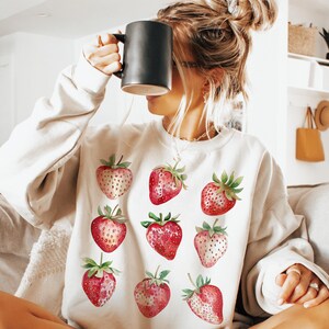 Op de afbeelding: Een wit sweatshirt met een rode aardbeienprint. De aardbeien zijn in een rasterpatroon gerangschikt en hebben groene bladeren.