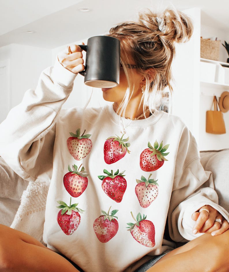 Op de afbeelding: Een wit sweatshirt met een rode aardbeienprint. De aardbeien zijn in een rasterpatroon gerangschikt en hebben groene bladeren.