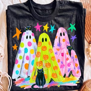 May include: A black t-shirt featuring a graphic of three ghosts and three black cats. The left ghost has colorful polka dots, the center ghost has orange pumpkins, and the right ghost has pink floral patterns. Brightly colored stars surround the ghosts.