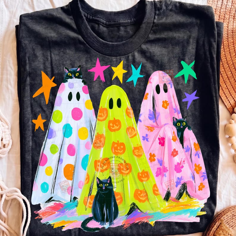 May include: A black t-shirt featuring a graphic of three ghosts and three black cats. The left ghost has colorful polka dots, the center ghost has orange pumpkins, and the right ghost has pink floral patterns. Brightly colored stars surround the ghosts.
