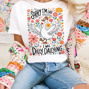 May include: White t-shirt with a floral design and a cartoon goose. The text on the shirt reads "Sorry I'm Late I Was Dilly Dallying".