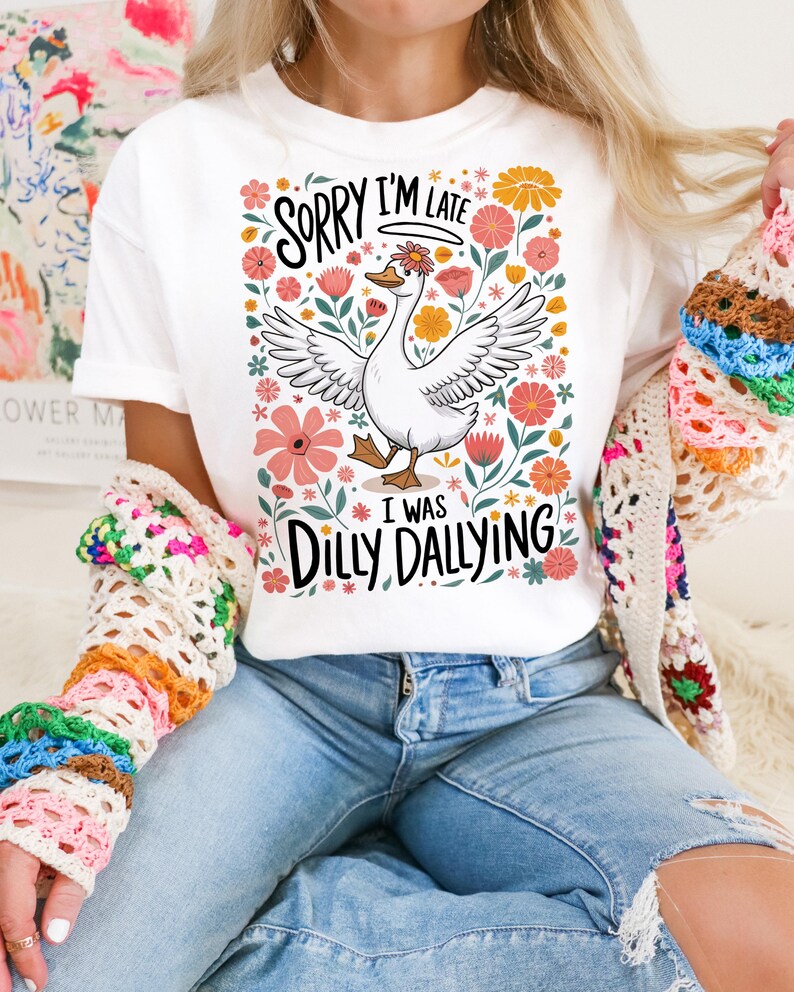 May include: White t-shirt with a floral design and a cartoon goose. The text on the shirt reads "Sorry I'm Late I Was Dilly Dallying".
