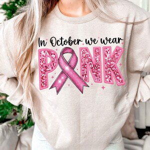 May include: A light beige sweatshirt with the text "In October we wear PINK" in pink glitter letters. A pink ribbon with glitter is in the center of the text.