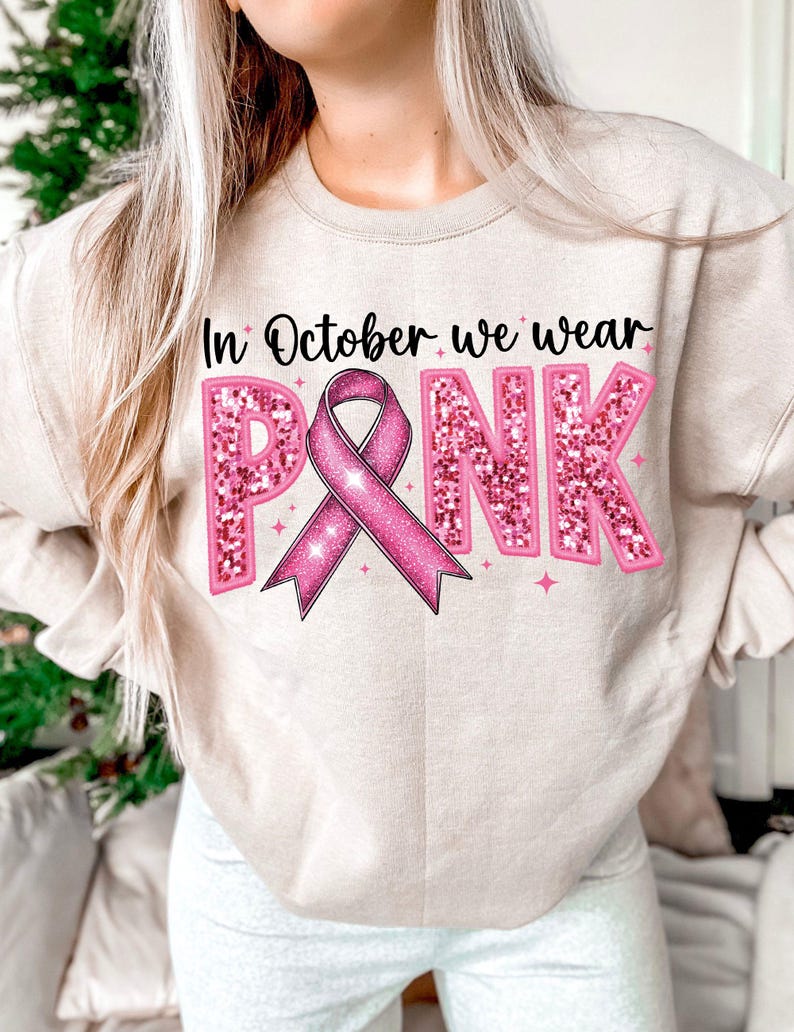 May include: A light beige sweatshirt with the text "In October we wear PINK" in pink glitter letters. A pink ribbon with glitter is in the center of the text.