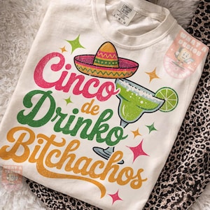 Peut inclure: T-shirt crème avec un motif coloré du Cinco de Mayo. Le graphisme comprend un sombrero, une margarita et les mots "Cinco de Drinko Bitchachos" en rose, vert et orange.