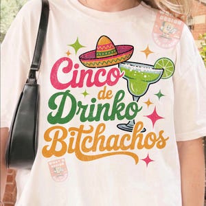 May include: Off-white t-shirt with a colorful Cinco de Mayo design. The graphic includes a sombrero, margarita, and the words "Cinco de Drinko Bitchachos" in various colors. A black shoulder bag is visible.