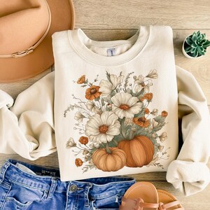 Pu&ograve; includere: Felpa color crema con un design floreale e zucca. La grafica include fiori bianchi e arancioni e due zucche. Un cappello color cachi e jeans blu sono anche nell'immagine, insieme a una piccola pianta succulenta.