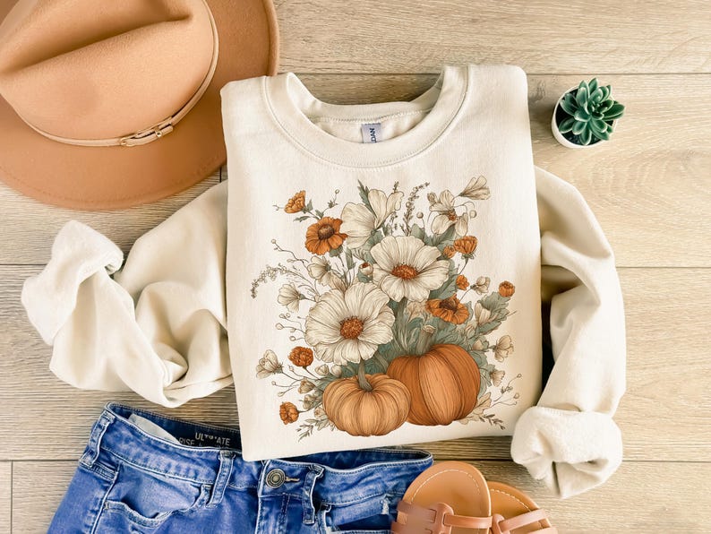 Pu&ograve; includere: Felpa color crema con un design floreale e zucca. La grafica include fiori bianchi e arancioni e due zucche. Un cappello color cachi e jeans blu sono anche nell'immagine, insieme a una piccola pianta succulenta.